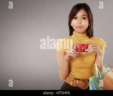 Gerne Asiatin mit vielen Einkaufstüten und Kreditkarte in der Hand vor grauem Hintergrund Stockfoto
