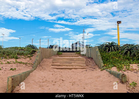 Holztreppen mit Pol Barriere neben Aloe-Pflanzen und Dünenvegetation gegen blauen Wolkenhimmel am Strandaufgang in Südafrika Stockfoto