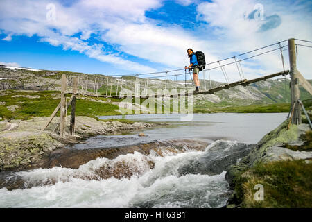 Junge Frau mit Rucksack wandern über schmale hölzerne Hängebrücke über den kleinen Fluss Stockfoto