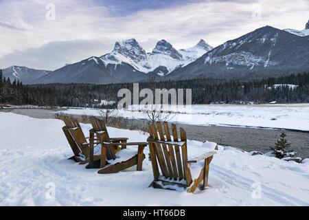 Wood Adirondack Stühle Bow River Bank Three Sisters Mountain Peak Aussichtspunkt. Kalter Wintertag Schneelandschaft Canmore Alberta Foothills Kanadische Rocky Mountains Stockfoto