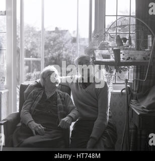 historische, der 1960er Jahre in den hinteren Raum ihres Hauses mit den französischen Türen offen zum Garten, ein glückliches paar ältere sitzen und studieren ihre Vögel im Käfig. Stockfoto