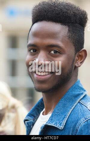 Ein Portrait eines jungen Mannes in die Kamera Lächeln. Er steht im freien trägt eine blaue Jeansjacke. Der Hintergrund ist unscharf. Stockfoto