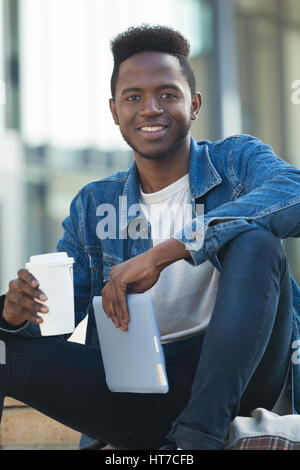 Eine glückliche junge Studentin lächelt in die Kamera, als er eine Auszeit vom Studium mit einem heißen Getränk nimmt. Er sitzt im Freien halten seine digital-Tablette. Stockfoto