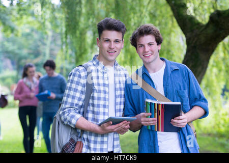Zwei glückliche männliche Studenten Lächeln, als sie im Freien und Studie zu entspannen. Sie halten ihre Taschen, Bücher und ein digital-Tablette, wie sie in die Kamera blickt. Stockfoto