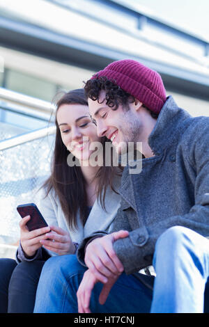 Ein glückliches junges Paar Lächeln, als sie im Freien entspannen. Sie nutzen ein Handy beim Sitzen auf einige Stadt-Schritte gemeinsam lachen. Stockfoto