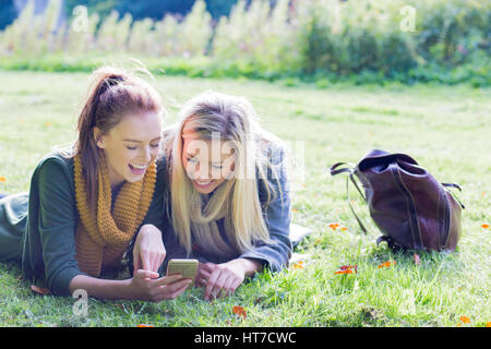 Zwei glückliche junge Studentinnen Lachen auf ihrem Handy, als sie eine Pause im Freien genießen. Sie sind einige Gras liegend. Stockfoto