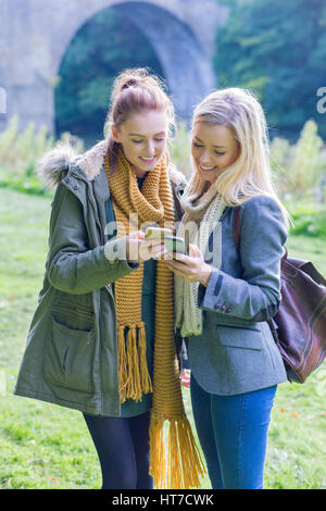 Zwei Studentinnen glückliche Lächeln, als sie ihre Mobiltelefone im freien gemeinsam nutzen. Sie sind warm stehen in der Nähe eines Flusses verpackt. Stockfoto