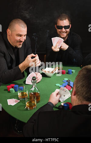 Foto von drei Männern Pokern, Zigarren rauchen, trinken Whisky und einer spiegeln den Vogel an den Spieler mit der gute Pokerhand. Stockfoto