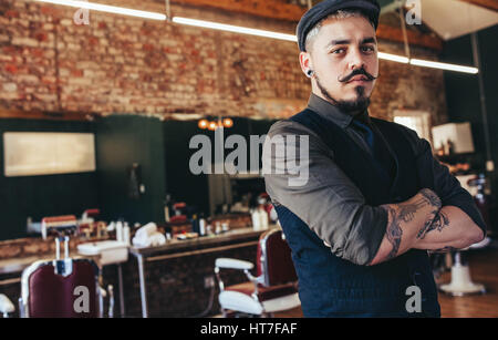 Porträt des jungen Mann, der mit seinen Armen kreuzten sich im Friseurladen. Stilvolle Friseur in seinem Salon Blick in die Kamera ernsthaft. Stockfoto