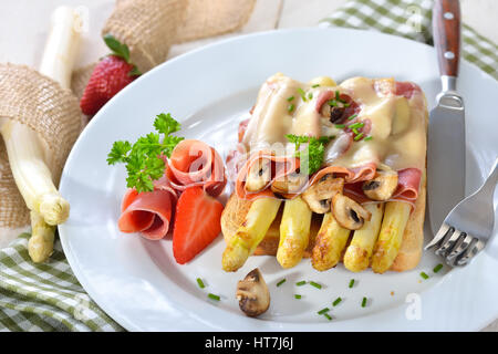 Leckeren Toast mit gebratenem weißen Spargel, spanischen Serrano Schinken, Champignons und Käse überbacken, serviert auf einem weißen Teller mit Erdbeeren Stockfoto