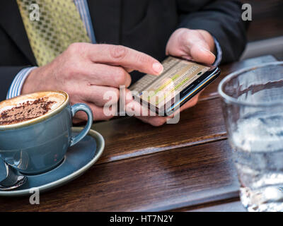 Geschäftsmann texting Die ausgereifte Geschäftsmann Hände schließen Texting mit Smartphone bei Alfresco Restaurant mit einem Glas Wasser und Cappuccino sitzen Stockfoto