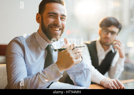 Porträt der jungen attraktiven Firmenleiterin mit gefalteten Händen am Tisch sitzen, halten Sie Stift und fröhlich in die Kamera Lächeln, sitzt neben seinem Stockfoto