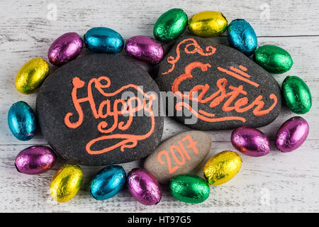 Glücklich Ostern 2017 Schriftzug auf Kieselsteinen geschrieben mit Schokoladeneier in bunter Folie auf weißem Holz Hintergrund verpackt Stockfoto