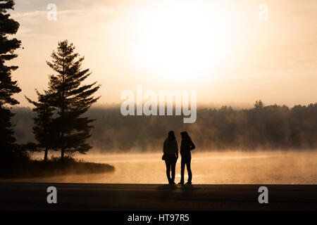 Zwei junge Frauen, die in ihrer Umgebung auf Nebel am frühen Morgen. See von zwei Flüssen, Algonquin Provincial Park, Ontario, Kanada. Stockfoto