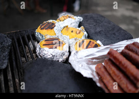 Traditionelle rumänische gegrillte Polenta mit Käse genannt "Bulz" in Folie und gegrillten Würstchen auf dem Grill in Transfagarasan Highway, Rumänien. Stockfoto