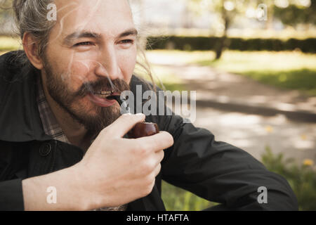 Junge asiatische bärtiger Mann rauchen Pfeife im Sommerpark, lächelnd Nahaufnahme portrait Stockfoto