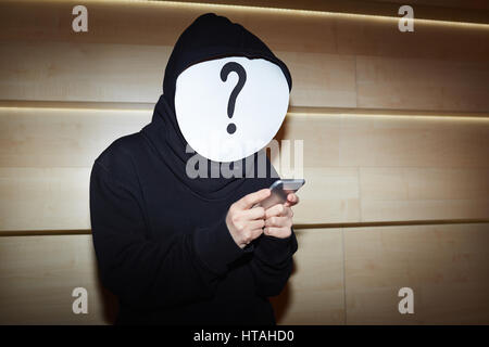 Hacker in schwarzer Hoodie und Fragezeichen Maske mit Smartphone zum Datendiebstahl stehend gegen Holzwand Stockfoto