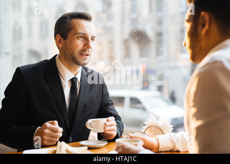 Business-Meeting im Café im warmen Abendlicht: zwei Geschäftsleute sprechen Angebote über Kaffee, konzentrieren sich auf stilvolle Mann mittleren Alters suchen überzeugend Stockfoto