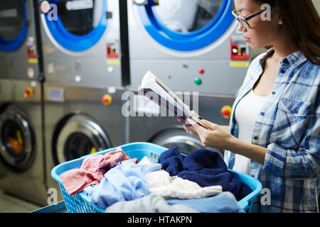 Junge Frau im Waschsalon mit Korb voller Kleidung lesen Stockfoto