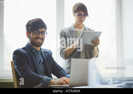 Porträt des erfolgreichen nahöstlichen Jungunternehmer mit Blick in die Kamera und lächelnd während der Arbeit mit Laptop, seine junge Sekretärin ich Brille Stockfoto
