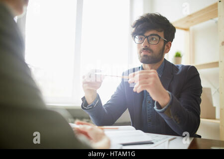 Porträt des jungen kreativen nahöstlichen Geschäftsmann Brille lauschte seinem Partner sitzt ihm gegenüber an Schreibtisch in sonnendurchfluteten modernen Stockfoto