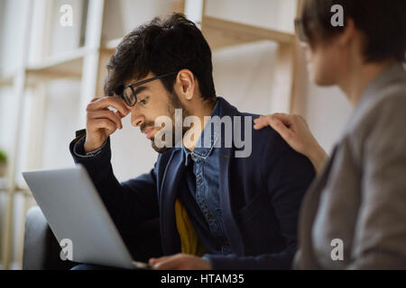 Seite Ansicht Porträt Exhaustd jungen nahöstlichen Geschäftsmann kneifen Nasensteg Blick auf Laptop-Bildschirm sitzen mit Kollegin helfen h Stockfoto