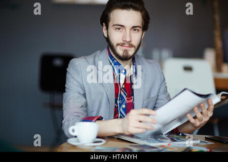Porträt des jungen langhaarigen Mann mit Business casual Kleidung sitzt am Tisch, Blick in die Kamera und lächelnd beim Lesen von Zeitschriften Stockfoto