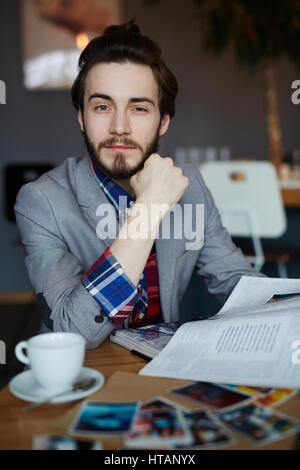 Porträt von zuversichtlich langhaarigen Mann mit Business casual Kleidung sitzt am Tisch, Blick in die Kamera und lächelnd beim Lesen von Zeitschriften Stockfoto