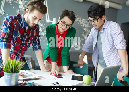 Gruppe von jungen kreativen Menschen tragen legere Kleidung Zusammenarbeit bei Treffen im Büro stehen Tisch und betrachten von Fotos und Dokumenten Stockfoto