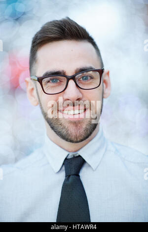 Kopf und Schultern Porträt der jungen zuversichtlich bärtigen Geschäftsmann tragen Brillen und weißen Hemd mit Krawatte gegen unscharfe Büro fröhlich lächelnd Stockfoto