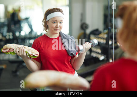 Portrait von junge übergewichtige Frau tun Gewicht Übungen mit riesigen dicken Sandwich beim Training im Fitness-Studio, Kampf der Diät, Gewicht zu verlieren Stockfoto