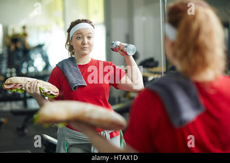 Porträt von niedlichen übergewichtige Frau mit riesigen dicken Sandwich und Hantel Übungen bestimmt, Gewicht zu verlieren, beim Training im Fitness-Studio Stockfoto