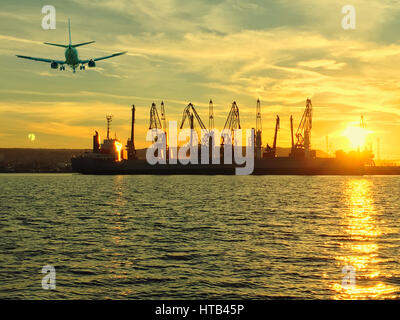 Flugzeug und Silhouetten von Kränen und industrielle Frachtschiffe im Hafen bei Sonnenuntergang Stockfoto