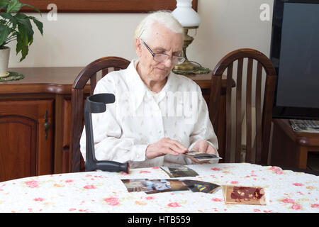 Alte Frau sitzt im Wohnzimmer am Tisch und schaut alte Fotografien. Alte Großmutter beobachten alte Fotos, die am Tisch sitzen. Stockfoto
