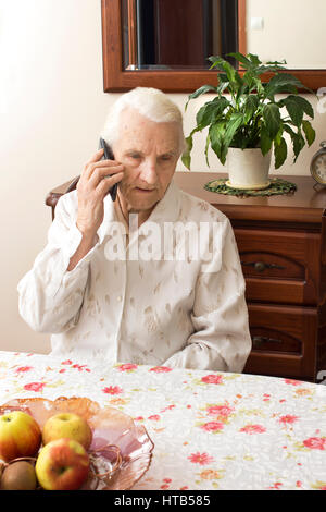 Großmutter sitzt an einem Tisch im Wohnzimmer und hält in seiner Hand das Telefon. Die alte Dame auf einem Handy beim Sitzen an einem Tisch im Gespräch Stockfoto