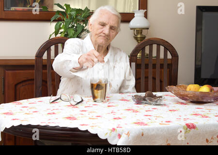 Die alte Frau sitzt an einem Tisch im Wohnzimmer und in einem Glas Tee zu machen. Stockfoto
