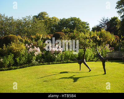 Statuen auf dem Rasen im Garten Chenies Manor, Buckinghamshire mit Sitzbank Sitz, rosa krautige Pflanzen, Sträucher, Rosen, Bäume und blauer Himmel. Stockfoto