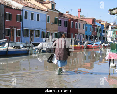 Farbenfrohe Burano-Kanäle spiegeln sich in Hochwasser (Aqua alta), während eine Frau bei Flut durch das Wasser geht Stockfoto