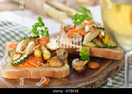 Vegetarischer Toast mit gebratenem weißen Spargel und anderes Gemüse auf einem Holzbrett serviert mit einem Glas Weißwein Stockfoto