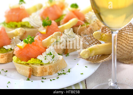 Köstliche Häppchen mit deutschen weißen Spargel, Frischkäse mit Kräutern, geräucherter Lachs auf italienische Ciabatta Brot mit Salatblättern, serviert mit einer Gla Stockfoto