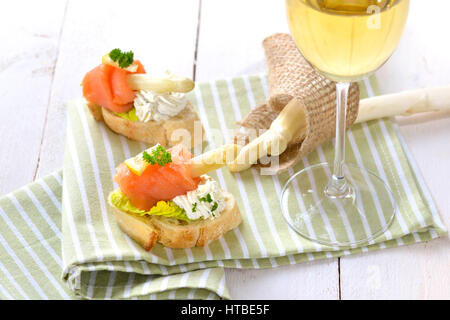 Köstliche Häppchen mit weißem Spargel, Frischkäse mit Kräutern, geräucherter Lachs auf italienische Ciabatta-Brot mit Salat, serviert mit einem Glas Wein Stockfoto