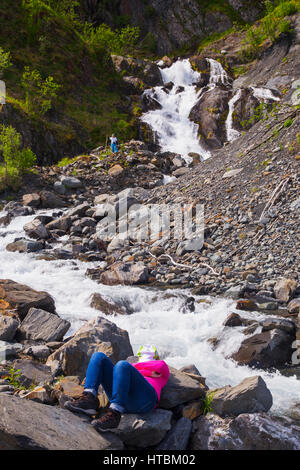 Eine Frau, die eine Pause von Seekajak in Kenai Fjords Nationalpark entspannt an einem rauschenden Bach, während ihre Partnerin einen Wasserfall erforscht Stockfoto