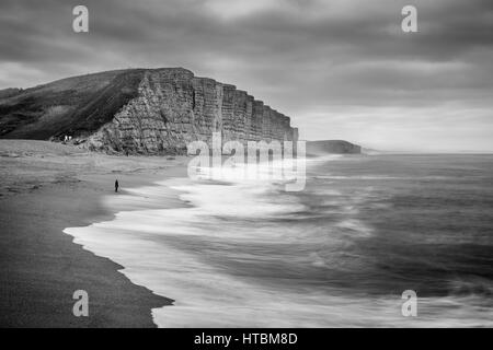 eine einsame Figur gerade Wellen brechen sich am Strand unter East Cliff, West Bay, Jurassic Coast, Dorset, England, UK Stockfoto