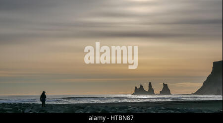 Silhouette der Soltiär Frau am Lavastrand in Vik, Island, in der Dämmerung mit Meeresstapeln und Klippen am niedrigen Horizont und launisch gelben Abend Winterhimmel Stockfoto