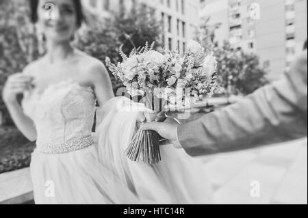 Braut und Bräutigam halten einen Blumenstrauß. Konzentrieren Sie sich auf das Bouquet. Frisch verheiratetes Paar Fuß auf Pfaden Händchenhalten und sahen einander. Gebäude in den Rücken Stockfoto