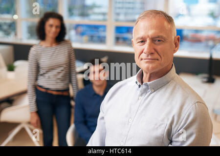 Porträt eines zuversichtlich und reifer Geschäftsmann stehend in einem modernen Büro mit zwei jungen Arbeitskollegen im Hintergrund Stockfoto