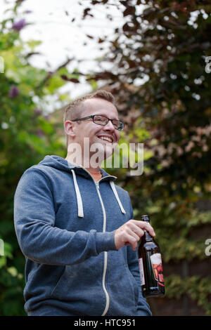Junge kurze blonde Haare Mann tragen Brillen und blauen Hoodie Pullover trinken Bier. Freunde treffen nach der Arbeit, im Garten grillen. Vertikale erschossen Stockfoto