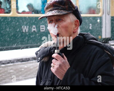 Alter Mann raucht eine Pfeife im Schnee am Bahnhof Wengen in der Schweiz. Ein Zug der Wengernalpbahn (WAB) ist im Hintergrund. Stockfoto