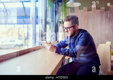 Ein freier Hipster-Mann mit Tablet in der Hand arbeitet Café am Fenster. Stockfoto