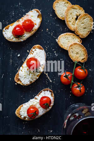 Toast mit weißen Schmelzkäse und gebratenen Kirschtomaten und Glas Rotwein bei schwarzem Hintergrund. Ansicht von oben Stockfoto